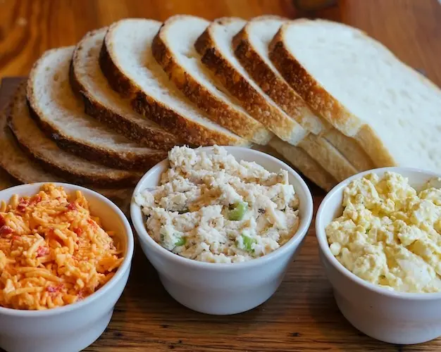 Three white bowls, each with a spread - Pimento Cheese, Chicken Salad, & Egg Salad - in front of several slices of sourdough breads.